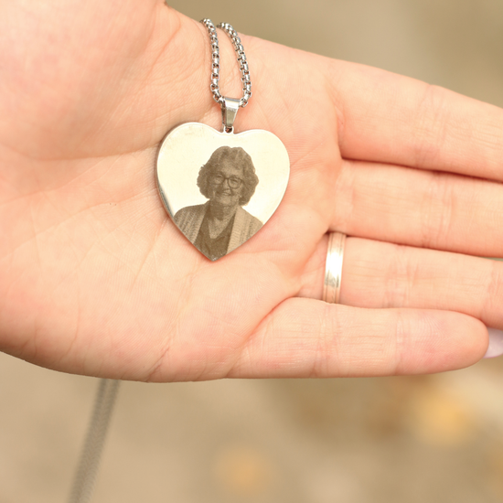 Un primer plano de una mano sosteniendo un collar con foto grabada en forma de corazón, que muestra el retrato sonriente de una mujer mayor. La mano luce un sencillo anillo en el dedo anular.