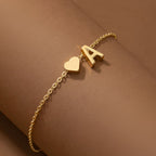 A Pulsera de tobillo con inicial en color dorado luce una fina cadena, un dije de corazón y una letra A, evocando elegancia sobre un fondo marrón suave.