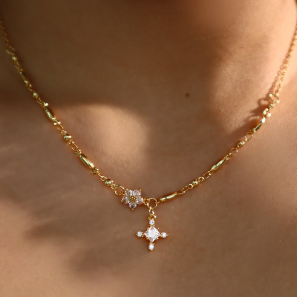 Primer plano del collar Destello de estrella en dorado, con un pequeño colgante en forma de flor con gemas diminutas y un colgante más grande en forma de diamante adornado con piedras brillantes, que se lleva alrededor del cuello.