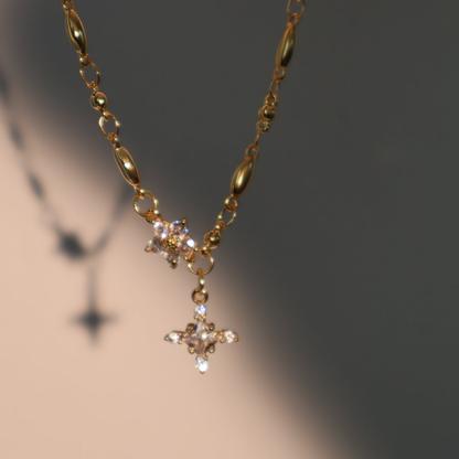 El Collar Destello de Estrella es un collar de oro con dos colgantes en forma de estrella, incrustados de gemas, que proyectan una suave sombra sobre un fondo beige y gris.