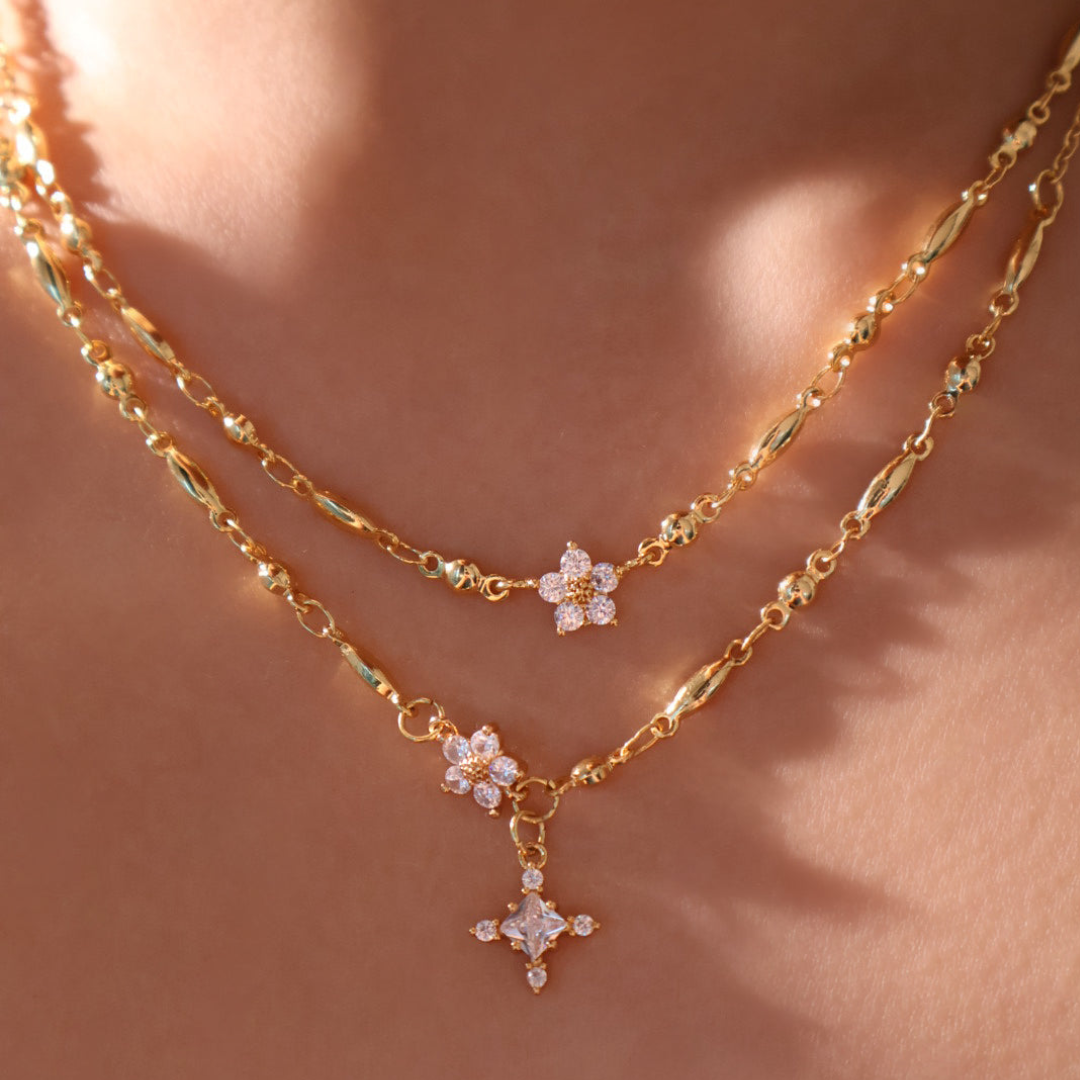 Dos collares de oro superpuestos en el cuello, cada uno con un delicado colgante floral de pequeñas gemas brillantes. El collar Destello de estrella presenta detalles de abalorios y texturas que reflejan maravillosamente la luz sobre la piel lisa.