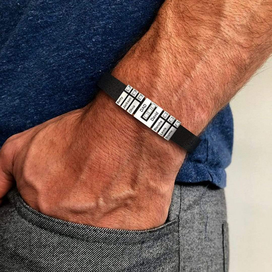La mano de una persona lleva una Pulsera de cuero personalizada con nombres, una pulsera negra con cuentas de plata grabadas, que descansa sobre unos pantalones grises. La persona lleva una camisa azul, un regalo único para papá.