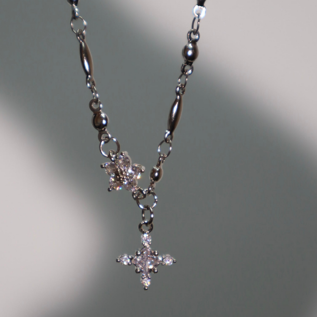 El collar Destello de estrella presenta dos colgantes en forma de estrella decorados con piedras claras, sobre un fondo gris claro suavemente iluminado.