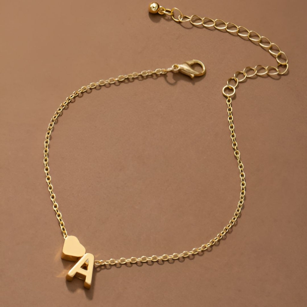 La Pulsera de tobillo con inicial, una delicada tobillera de oro con un pequeño dije de corazón y un dije de letra A, se muestra sobre un fondo marrón. Tiene una cadena ajustable con cierre de langosta y eslabones extensibles.