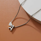 Un collar de plata con una A y un colgante de corazón descansa sobre una superficie lisa de color marrón junto a un objeto curvo blanco, emparejado con una Pulsera de tobillo con inicial para darle un toque personalizado.
