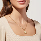 Una mujer lleva un top beige acanalado y el Collar Marco con inicial personalizada (Oferta 2x1), un colgante de oro con un dije rectangular ornamentado. La imagen está recortada para mostrar el cuello, el pecho y parte de la cara.