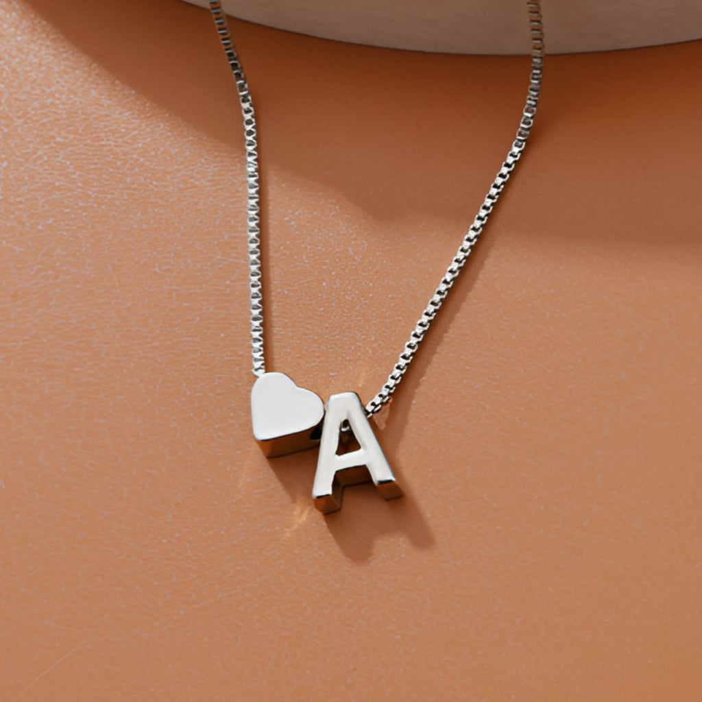 Un collar de plata con un charm en forma de corazón y un colgante con la letra A descansa sobre una superficie de color marrón claro, que combina a la perfección con la elegancia de la Pulsera de tobillo con inicial.