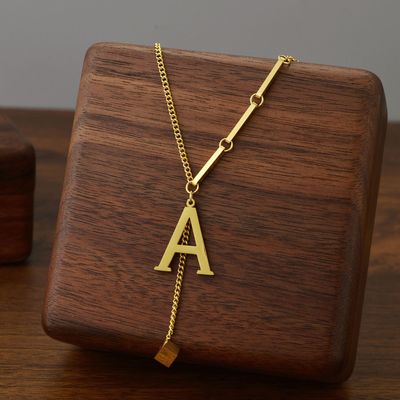 Collar Letra (Oferta 2x1): elegante collar de acero inoxidable con colgante de letra A y detalle geométrico, presentado sobre bloque de madera. ¡Incluye envío gratis!.