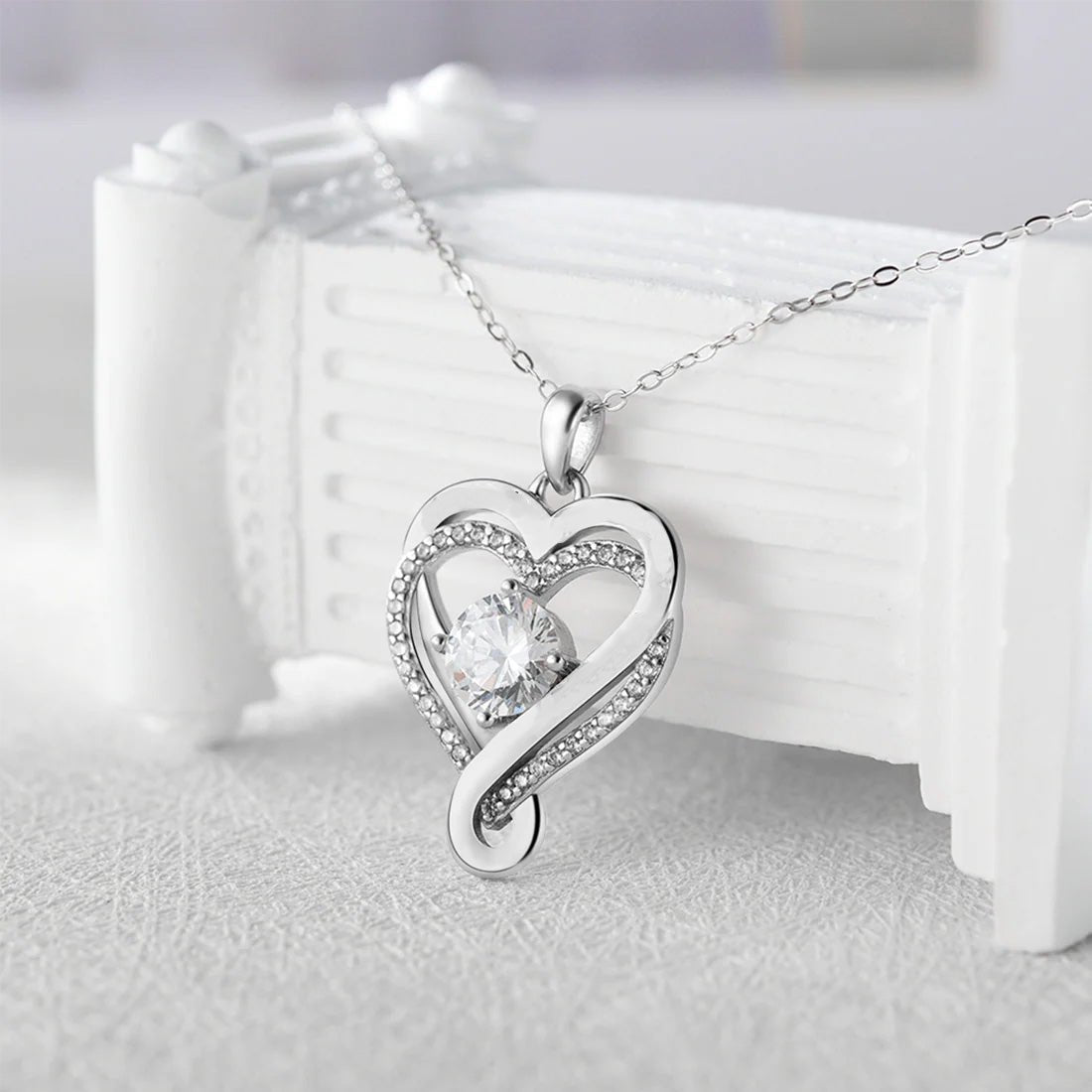 El "A mi esposa" - Collar corazón para siempre presenta un colgante de plata en forma de corazón con una brillante gema central y cristales a lo largo del contorno, elaborado en acero inoxidable, sobre una delicada cadena y desplegado sobre una superficie decorativa blanca.
