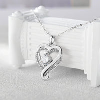 El "A mi esposa" - Collar corazón para siempre presenta un colgante de plata en forma de corazón con una brillante gema central y cristales a lo largo del contorno, elaborado en acero inoxidable, sobre una delicada cadena y desplegado sobre una superficie decorativa blanca.