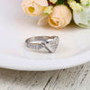 Un Anillo Abrazo al cielo de plata personalizado con pequeños diamantes se asienta sobre una placa blanca, delante de flores de color melocotón y blanco. El corazón lleva grabadas unas alas, un nombre y los años 1987-2022: un elegante anillo conmemorativo.