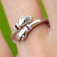 El Anillo Abrazo con piedras de nacimiento es un anillo de plata con forma de dos manos que envuelven el dedo, cada una de las cuales sostiene una piedra de nacimiento roja y otra morada, engastadas sobre un fondo verde.
