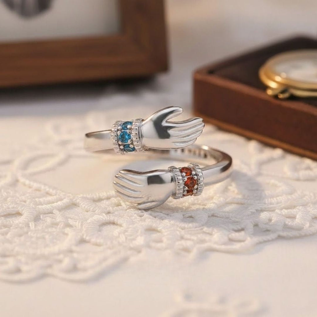 El Anillo Abrazo con piedras de nacimiento es un anillo de plata con dos manos enfrentadas, cada muñeca engastada con piedras de nacimiento azules y rojas. Se muestra apoyado sobre encaje blanco, con un reloj difuminado al fondo.