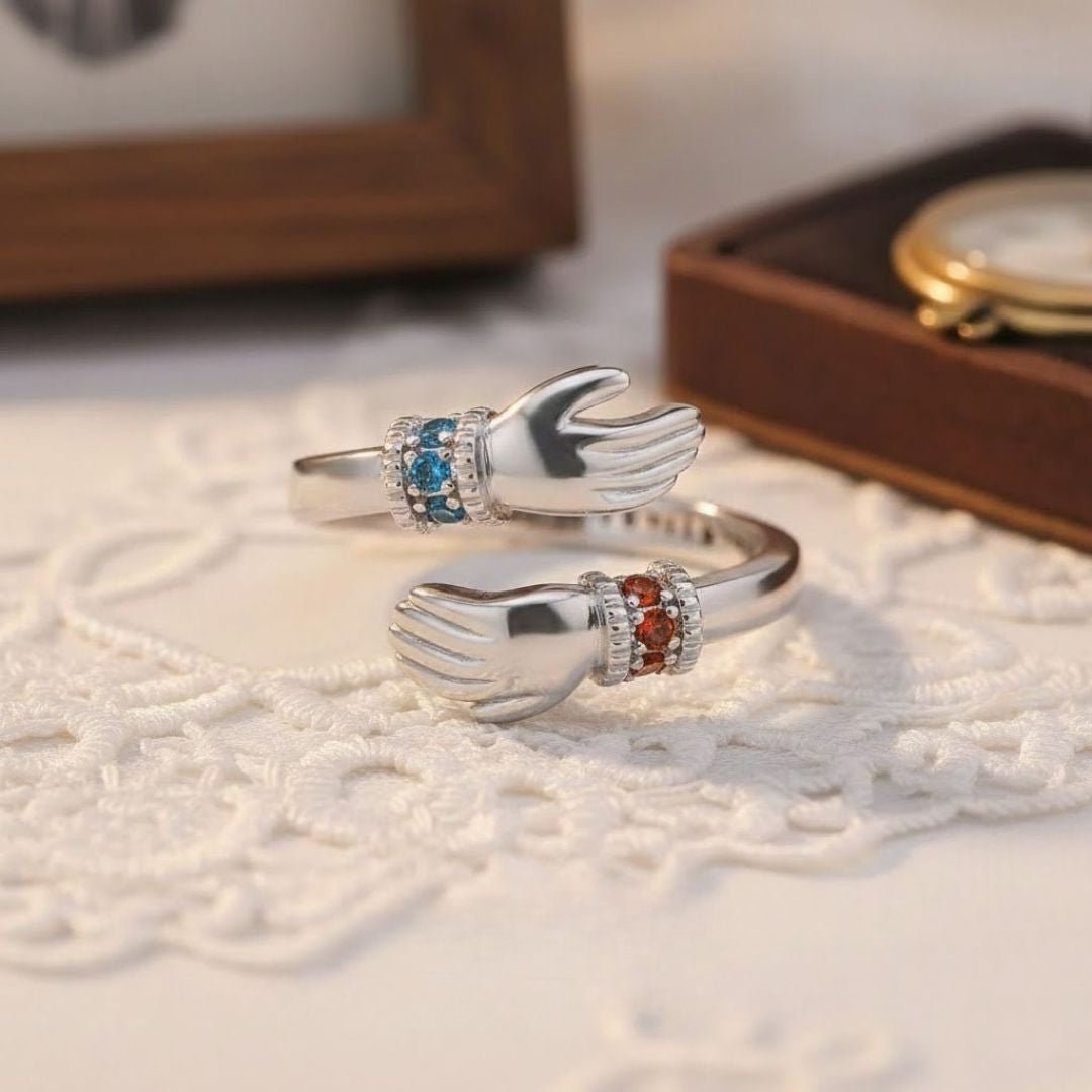 El Anillo Abrazo con piedras de nacimiento es un anillo de plata con dos manos enfrentadas, cada muñeca engastada con piedras de nacimiento azules y rojas. Se muestra apoyado sobre encaje blanco, con un reloj difuminado al fondo.