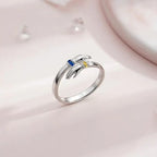 El Anillo Abrazo con piedras de nacimiento es un anillo de plata con dos manos que se tienden la una hacia la otra, cada una sosteniendo una piedra de nacimiento azul o amarilla, expuestas sobre una suave superficie rosa pálido.