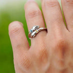 Primer plano de una mano que lleva el Anillo Abrazo con piedras de nacimiento, con dos pequeñas manos que sostienen piedras de nacimiento, sobre un fondo verde difuminado.