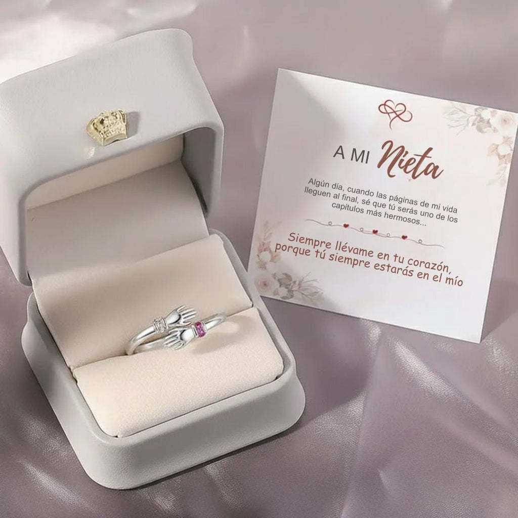 Un Anillo Abrazo con piedras de nacimiento, con dos manos sosteniendo un corazón, descansa en un joyero blanco junto a una tarjeta en español dirigida "A mi Nieta", que expresa que siempre estarán en el corazón del otro.