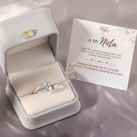 Un Anillo Abrazo con piedras de nacimiento, con dos manos sosteniendo un corazón, descansa en un joyero blanco junto a una tarjeta en español dirigida "A mi Nieta", que expresa que siempre estarán en el corazón del otro.