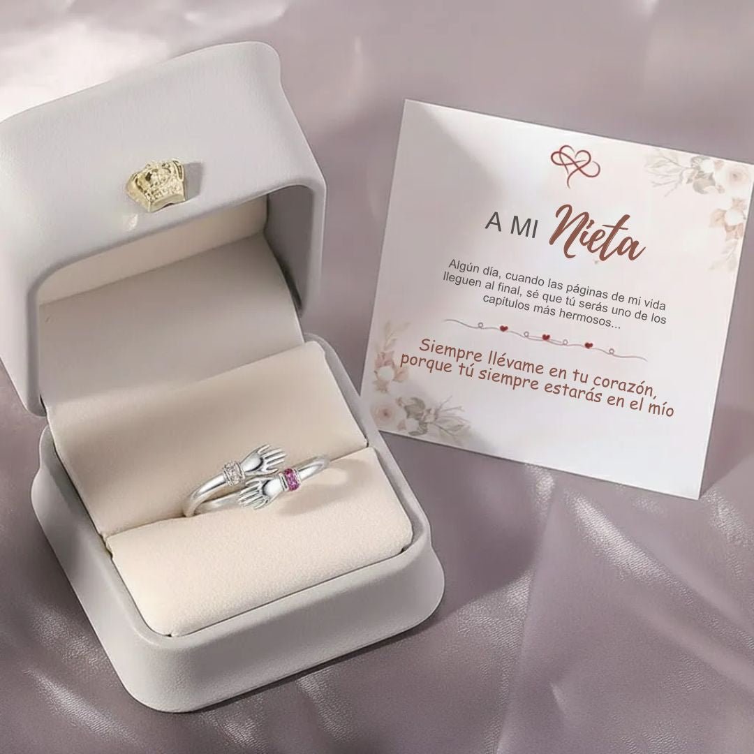 Un Anillo Abrazo con piedras de nacimiento, con dos manos sosteniendo un corazón, descansa en un joyero blanco junto a una tarjeta en español dirigida "A mi Nieta", que expresa que siempre estarán en el corazón del otro.