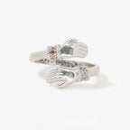 El Anillo Abrazo con piedras de nacimiento es de plata, con dos manos abiertas en los extremos, cada una luciendo una pulsera de gema diferente: una rosa y otra morada. Su diseño ajustable lo hace ideal como anillo personalizado.