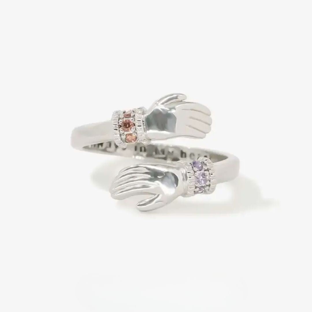 El Anillo Abrazo con piedras de nacimiento es de plata, con dos manos abiertas en los extremos, cada una luciendo una pulsera de gema diferente: una rosa y otra morada. Su diseño ajustable lo hace ideal como anillo personalizado.