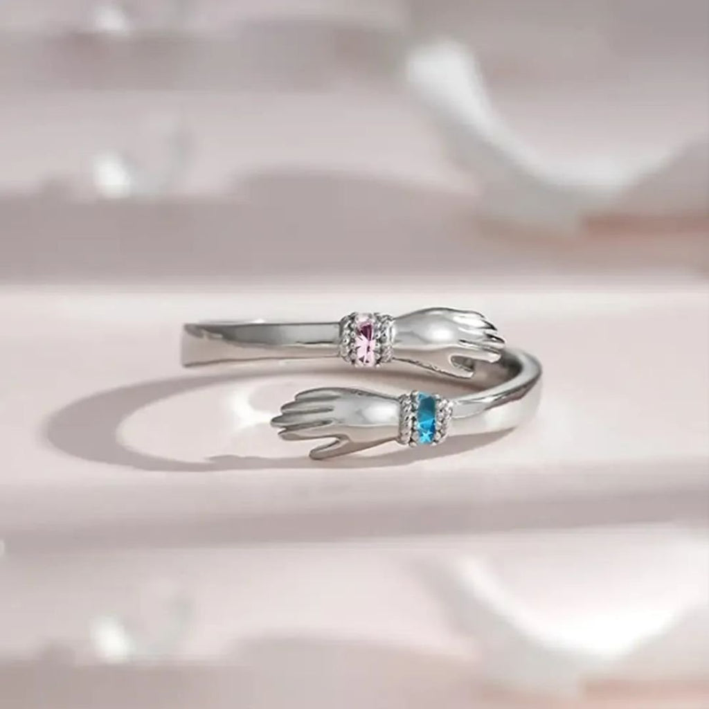 El Anillo Abrazo con piedras de nacimiento es un anillo plateado en forma de dos manos que se acercan, una decorada con una gema rosa y la otra con una piedra de nacimiento azul, mostradas sobre un fondo claro y suave.