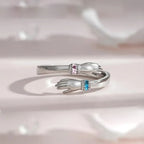 El Anillo Abrazo con piedras de nacimiento es un anillo plateado en forma de dos manos que se acercan, una decorada con una gema rosa y la otra con una piedra de nacimiento azul, mostradas sobre un fondo claro y suave.