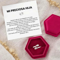 El Anillo Abrazo - "Mi preciosa hija", un anillo de plata en una caja hexagonal rosa con una tarjeta en español que expresa el amor y el apoyo de los padres, constituye un sentido regalo para hija, expuesto en una suave superficie decorativa de color claro.