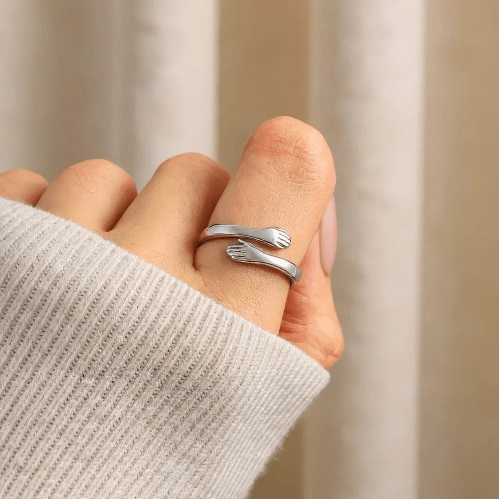 Una mano luce el Anillo Abrazo - "Mi preciosa hija", en forma de dos brazos plateados abrazando el dedo, con manos pequeñas en los extremos. Combinando estilo y significado, es un regalo perfecto para tu hija.