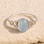 El Anillo ajustable Cristal de Mar presenta una pulsera de plata con una gema azul claro de forma ovalada, acentuada por delicados diseños de corazones, presentada sobre una superficie beige texturizada.