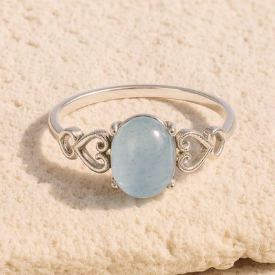 El Anillo ajustable Cristal de Mar presenta una pulsera de plata con una gema azul claro de forma ovalada, acentuada por delicados diseños de corazones, presentada sobre una superficie beige texturizada.