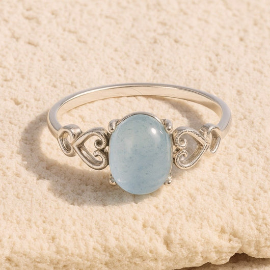 El Anillo ajustable Cristal de Mar presenta una pulsera de plata con una gema azul claro de forma ovalada, acentuada por delicados diseños de corazones, presentada sobre una superficie beige texturizada.