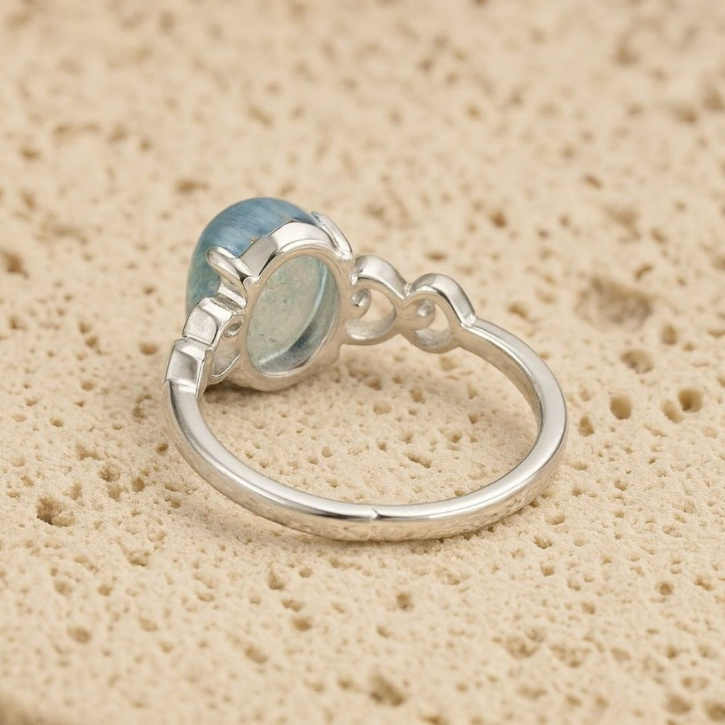 El Anillo ajustable Cristal de Mar presenta una gran gema ovalada de aguamarina engastada en un bisel decorativo sobre una banda de plata lisa, desplegada sobre una superficie de textura beige.