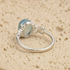 El Anillo ajustable Cristal de Mar presenta una gran gema ovalada de aguamarina engastada en un bisel decorativo sobre una banda de plata lisa, desplegada sobre una superficie de textura beige.