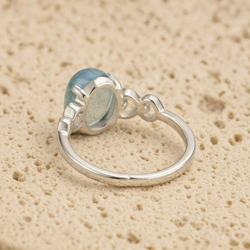El Anillo ajustable Cristal de Mar presenta una gran gema ovalada de aguamarina engastada en un bisel decorativo sobre una banda de plata lisa, desplegada sobre una superficie de textura beige.