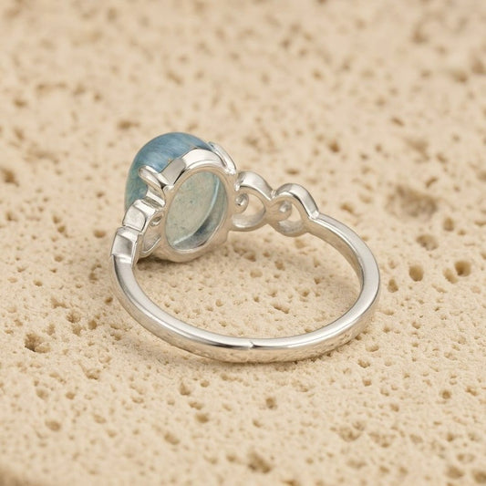 El Anillo ajustable Cristal de Mar presenta una gran gema ovalada de aguamarina engastada en un bisel decorativo sobre una banda de plata lisa, desplegada sobre una superficie de textura beige.