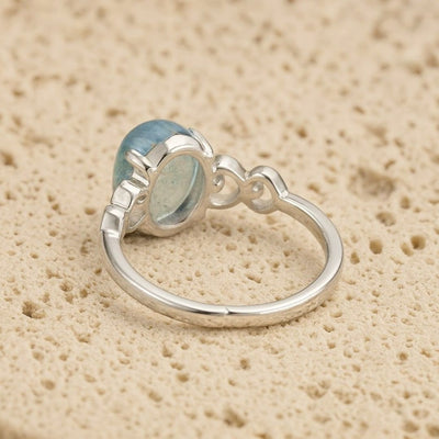 El Anillo ajustable Cristal de Mar presenta una gran gema ovalada de aguamarina engastada en un bisel decorativo sobre una banda de plata lisa, desplegada sobre una superficie de textura beige.