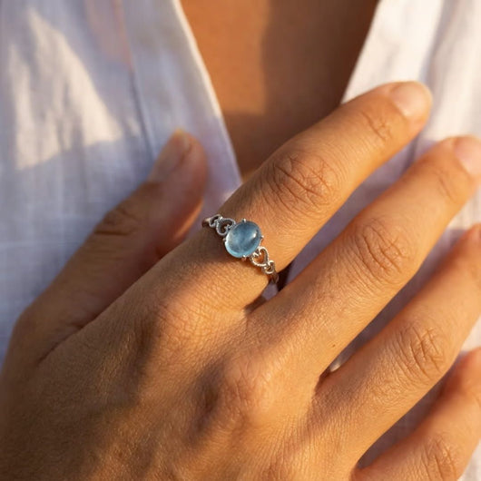 Una mano de tez morena clara luce el Anillo ajustable Cristal de Mar, con una llamativa gema azul, que descansa suavemente sobre una camisa blanca parcialmente abierta.