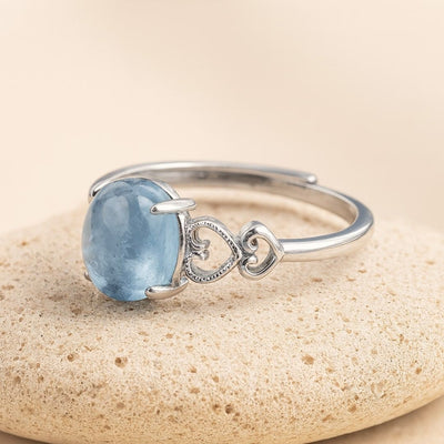 El Anillo ajustable Cristal de Mar presenta una gran gema ovalada de aguamarina y dos diseños de corazones entrelazados en una pulsera de plata, sobre un fondo de piedra beige claro.