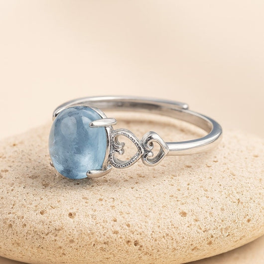 El Anillo ajustable Cristal de Mar presenta una gran gema ovalada de aguamarina y dos diseños de corazones entrelazados en una pulsera de plata, sobre un fondo de piedra beige claro.