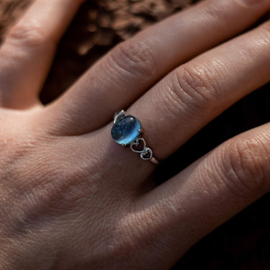 Primer plano de una mano que lleva el Anillo ajustable Cristal de Mar, con una gema ovalada de aguamarina azul y un pequeño corazón de plata en la correa, sobre un fondo marrón texturizado.