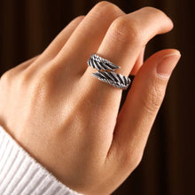 Cargar imagen en el visor de la galería, Primer plano de una mano de piel clara que lleva el Anillo Alas de Ángel, un anillo de plata con forma de dos alas de ángel superpuestas con detallados dibujos de plumas, combinado con un top blanco de manga larga.