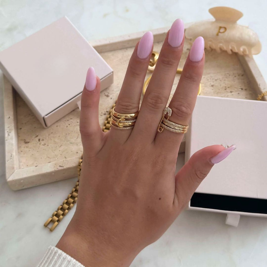 Una mano con largas uñas rosa pastel lleva varios anillos de oro y diamantes, entre ellos el Anillo apilable brillante. Al fondo hay piezas de joyería doradas, una bandeja de joyas beige, una pinza para el pelo y cajas rosas sobre una superficie de mármol.
