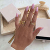 Una mano con largas uñas rosa pastel lleva varios anillos de oro y diamantes, entre ellos el Anillo apilable brillante. Al fondo hay piezas de joyería doradas, una bandeja de joyas beige, una pinza para el pelo y cajas rosas sobre una superficie de mármol.