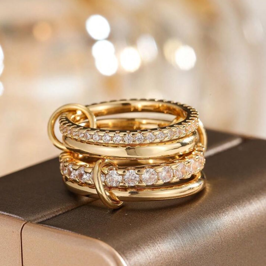 Primer plano de tres anillos Anillo apilable brillante de acero quirúrgico 316L, cada uno con pequeñas circonitas cúbicas transparentes, apilados sobre una superficie marrón metálica con un fondo suavemente difuminado de tonos cálidos.