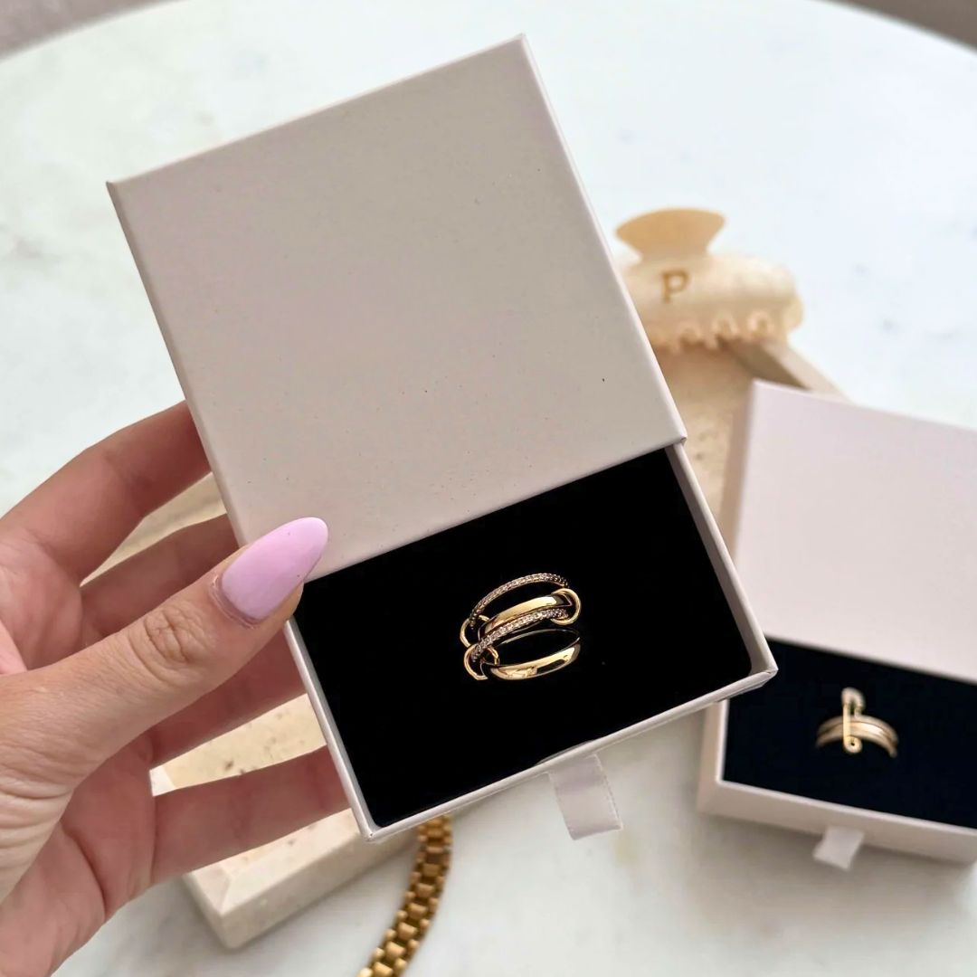 Una mano con las uñas pintadas de rosa claro sostiene un joyero beige abierto en el que aparece el Anillo apilable brillante en oro. Otra caja similar con un anillo de oro aparece al fondo sobre una superficie blanca.