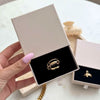 Una mano con las uñas pintadas de rosa claro sostiene un joyero beige abierto en el que aparece el Anillo apilable brillante en oro. Otra caja similar con un anillo de oro aparece al fondo sobre una superficie blanca.