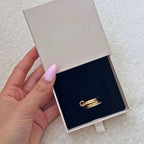 Una mano con las uñas pulidas de color rosa claro sostiene un joyero beige abierto que contiene un Anillo apilable brillante, expuesto en un interior de terciopelo negro.