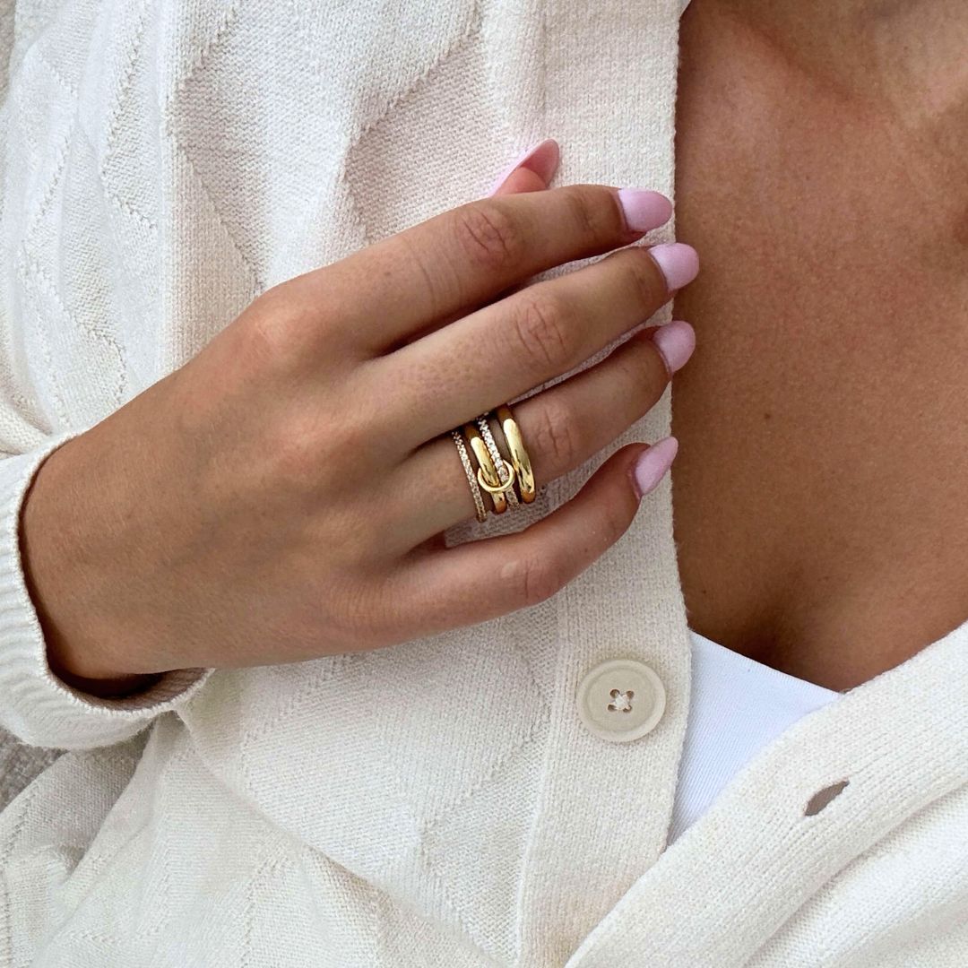 Una persona con las uñas cuidadas de color rosa claro lleva varios anillos de oro, con el Anillo apilable brillante. Lleva una rebeca color crema sobre un top blanco y apoya la mano cerca del pecho para mostrar tanto las joyas como el atuendo.