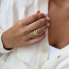 Una persona con las uñas cuidadas de color rosa claro lleva varios anillos de oro, con el Anillo apilable brillante. Lleva una rebeca color crema sobre un top blanco y apoya la mano cerca del pecho para mostrar tanto las joyas como el atuendo.